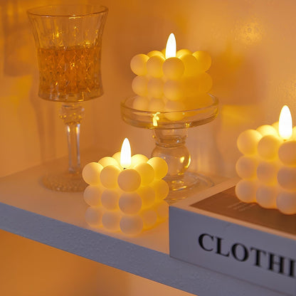 Drei LED-Kerzen in Würfelform, warmweißes Licht, auf Regal neben Kristallglas und Buch.